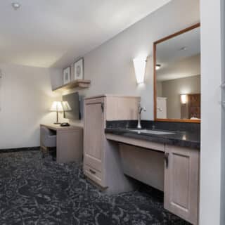 Accessible hotel room with wide entry, lowered sink, large mirror, workspace with lamp and TV, patterned carpet, and window with cityscape view.