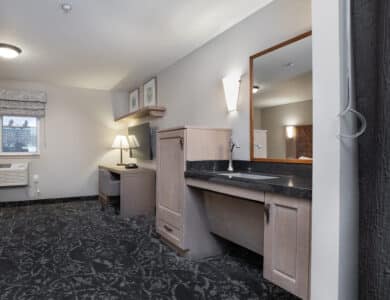 Accessible hotel room with patterned carpet, desk area, wall-mounted TV, window with air conditioner, and a sink with space underneath for wheelchair access.