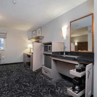 Hotel room with a workspace, window, air conditioner, and kitchenette area featuring a sink, microwave, coffee maker, and cabinets with pull-out shelves.