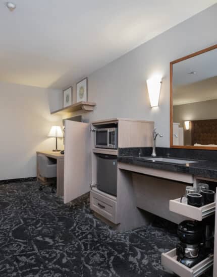 Hotel room kitchenette with microwave, sink, and pull-out drawers; work desk with lamp and chair by the window; neutral-colored walls and carpeted floor.