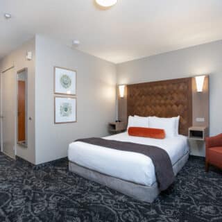 A hotel room with a double bed, two wall lamps, framed artwork, an orange chair, a wooden headboard, and a door leading to the hallway.