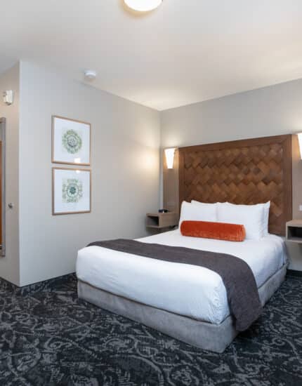 A hotel room with a double bed, two wall lamps, framed artwork, an orange chair, a wooden headboard, and a door leading to the hallway.