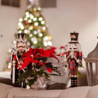 Two nutcracker figurines and a clear vase with red poinsettias are displayed on a table; a blurred Christmas tree with lights is in the background.
