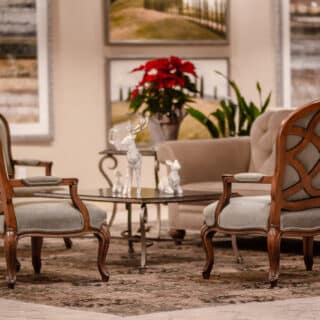 A seating area with two wooden armchairs, a glass coffee table, a beige sofa, potted red flowers, and framed landscape art on the wall.