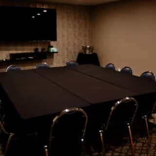 A small conference room with a rectangular table covered in black cloth, eight chairs, a wall-mounted TV, and some items on a shelf and side table.