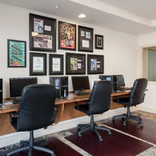 A row of four desktop computers with black chairs in a well-lit office space; framed certificates and photos are displayed on the wall above the desks.