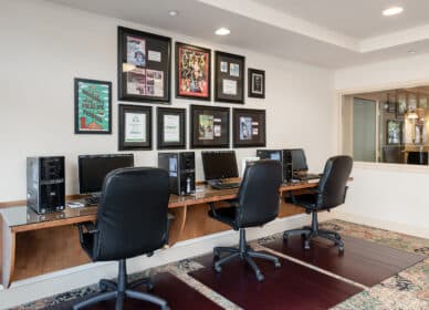 A row of four desktop computers with black chairs in a well-lit office space; framed certificates and photos are displayed on the wall above the desks.