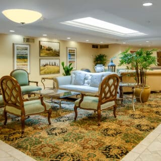 A brightly lit lobby with armchairs, sofas, glass coffee tables, potted plants, framed landscape art, and a patterned carpet on a tiled floor.