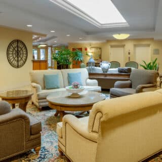 A hotel lobby with cushioned sofas and chairs arranged around coffee tables, decorative plants, a patterned rug, and soft lighting.