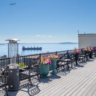 Rooftop patio with tables, chairs, and planters overlooks a body of water with ships and an airplane in the clear blue sky.