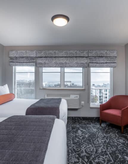 Hotel room with two beds, a window with a city and water view, a red chair, desk, lamp, and neutral-toned decor.