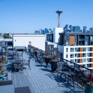 Rooftop patio with metal tables and chairs, potted plants along the railing, and a view of the Seattle skyline and Space Needle under a clear blue sky.