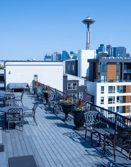 Rooftop patio with metal tables and chairs, potted plants along the railing, and a view of the Seattle skyline and Space Needle under a clear blue sky.