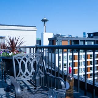 Rooftop patio with metal chairs, a potted plant, wine bottle, and glasses; city buildings and the Space Needle are visible in the background.
