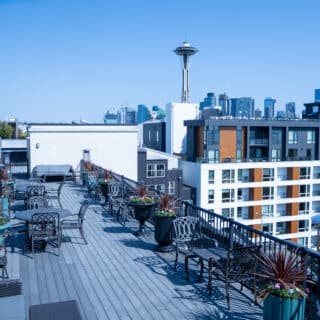 Rooftop patio with tables, chairs, and potted plants overlooking Seattle skyline, with the Space Needle visible in the background on a clear day.