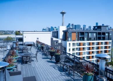 Rooftop patio with tables, chairs, and potted plants overlooking Seattle skyline, with the Space Needle visible in the background on a clear day.