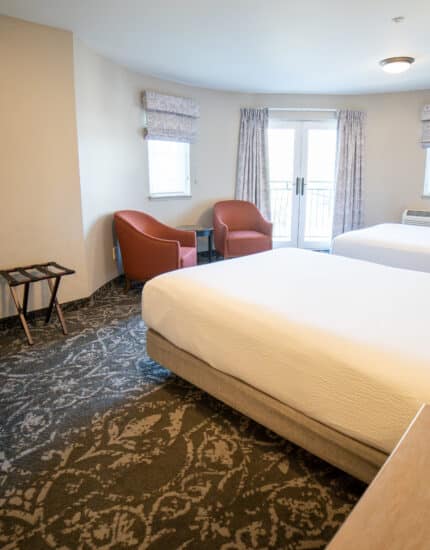 Hotel room with two double beds, a desk, two red chairs, a TV, and windows letting in natural light. The room has patterned carpet and neutral-colored walls.