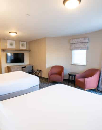 A hotel room with two beds, a TV, a small table with two chairs, a desk, and a window with curtains next to a door leading outside.