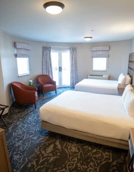 Hotel room with two double beds, a bedside lamp, two red chairs by the window, a TV on a dresser, phone, and patterned carpet. Natural light enters through large windows.