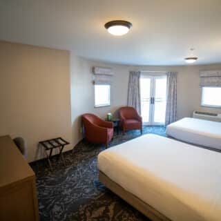 Hotel room with two double beds, a TV, two red chairs, a small table, dresser, and windows with curtains. The room is well-lit and has neutral-colored walls and carpeting.