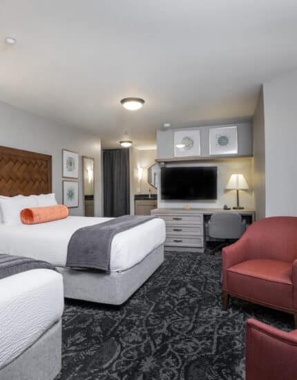 Hotel room with two queen beds, a TV on a dresser, a desk, two red chairs, a round table, and bedside lamps, decorated in neutral tones with framed art on the walls.