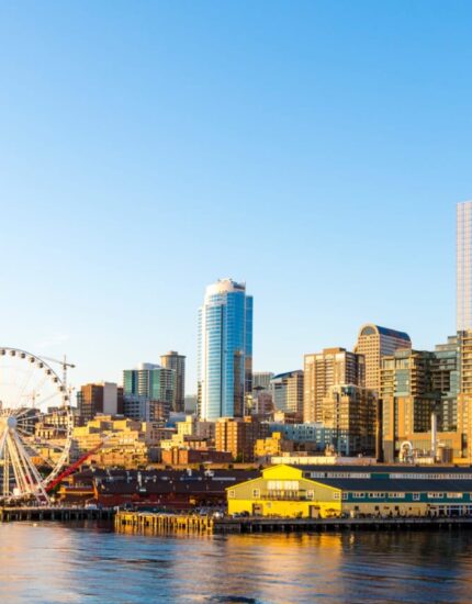 The Seattle waterfront skyline with the Ferris wheel, tall buildings, and boats docked along the water, viewed on a clear, sunny day.