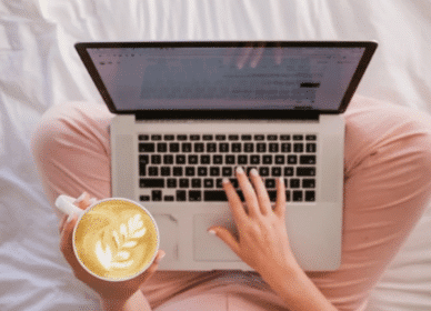 Person sitting cross-legged on a bed, using a laptop with one hand and holding a cup of latte with latte art in the other hand.