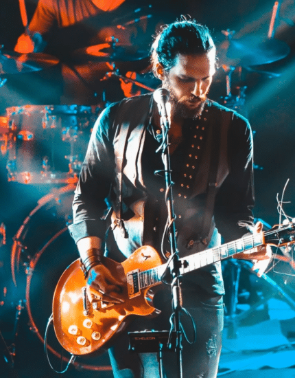 A musician plays an electric guitar on stage under blue and orange lights, with a drum set and other instruments visible in the background.