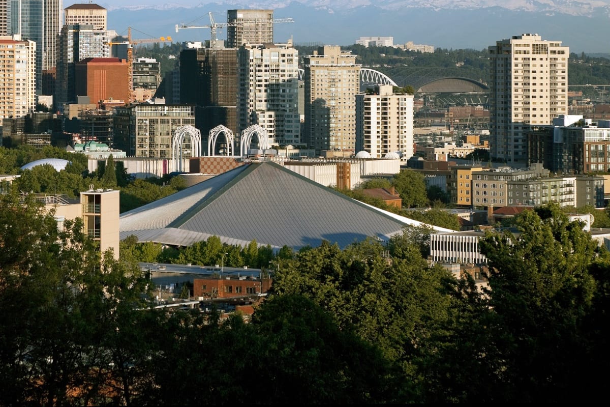 A large, pyramid-shaped arena surrounded by trees is set against a backdrop of tall city buildings and distant mountains.