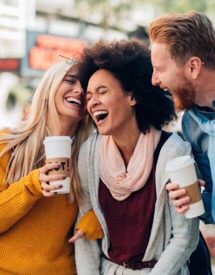 Five people stand close together outdoors, smiling and laughing while holding coffee cups, appearing to enjoy each other's company.