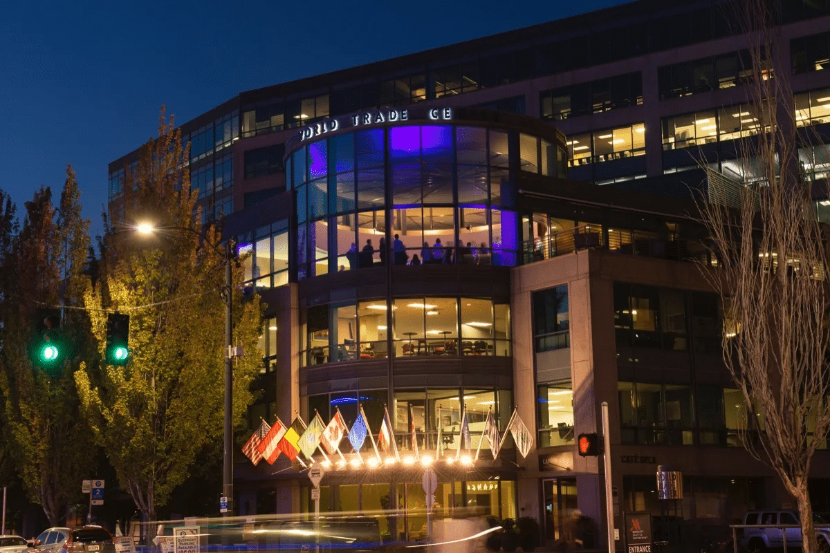 Modern office building at night with illuminated windows and several flags displayed at the entrance, labeled "World Trade Center." Trees and traffic lights flank the scene.