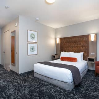 Hotel room with a double bed, white linens, an orange accent pillow, wall-mounted lights, patterned carpet, an armchair, a mirror, and framed art on the wall.