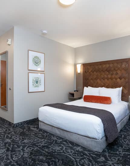 Hotel room with a double bed, white linens, an orange accent pillow, wall-mounted lights, patterned carpet, an armchair, a mirror, and framed art on the wall.