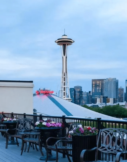 Rooftop patio with seating and outdoor heaters overlooking the Seattle skyline, including the Space Needle, under a cloudy sky.