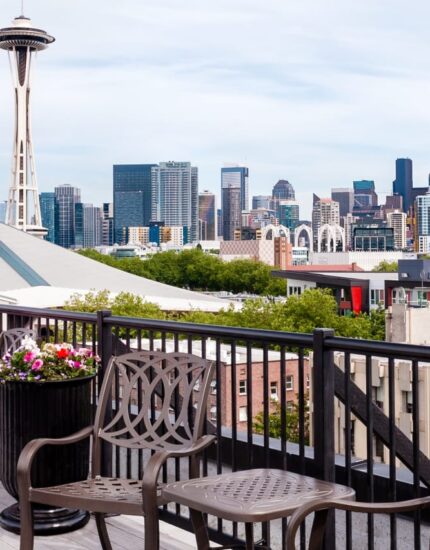 Rooftop deck with chairs and planters overlooks the Seattle skyline, featuring the Space Needle and downtown buildings under a partly cloudy sky.