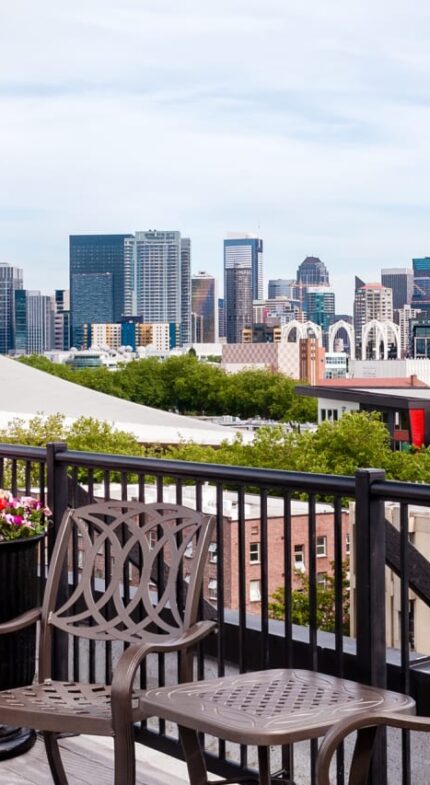 Rooftop deck with chairs and planters overlooks the Seattle skyline, featuring the Space Needle and downtown buildings under a partly cloudy sky.