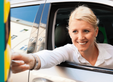 A woman in a white shirt is sitting in a car and reaching out through the window, smiling as she interacts with a device outside the vehicle.