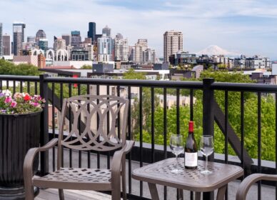 A metal patio chair and table with a wine bottle and two glasses overlook a city skyline with tall buildings, greenery, and a distant mountain.