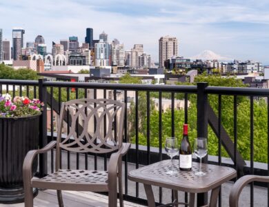 A metal patio chair and table with a wine bottle and two glasses overlook a city skyline with tall buildings, greenery, and a distant mountain.