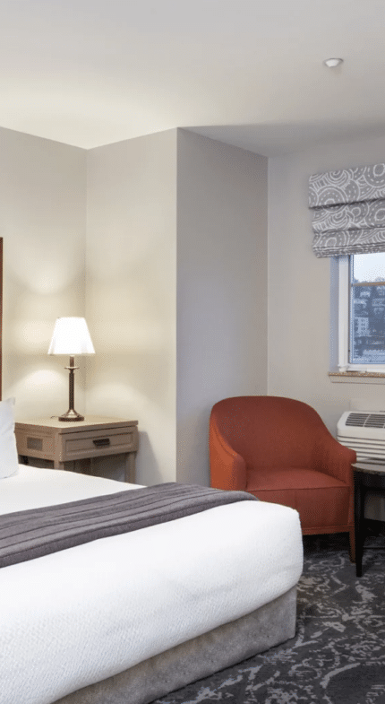 A hotel room with a king bed, two bedside tables with lamps, two red chairs by a window, and a desk on the right.