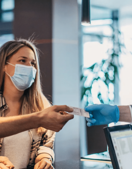 Two people wearing masks check in at a counter, handing a document to a staff member in a blue suit and mask.