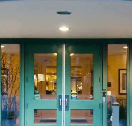 The entrance of a building with green double doors, large glass windows, two potted plants, and warm interior lighting visible inside.