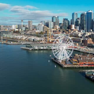 A large Ferris wheel stands on a waterfront pier in front of a city skyline with tall buildings and the Space Needle visible in the background.