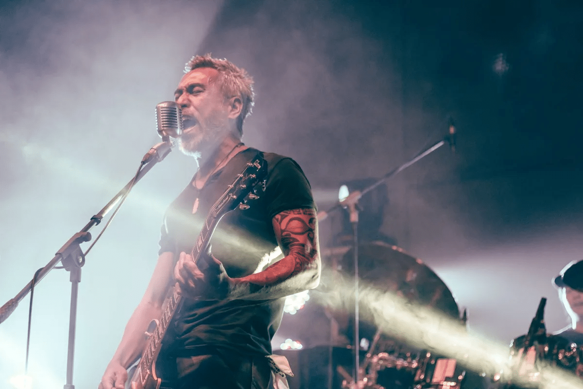 A musician with a guitar sings into a vintage microphone on stage, illuminated by stage lights, with a drummer playing in the background.