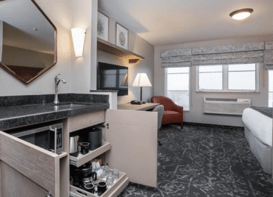 Hotel room with patterned carpet, a bed, a red armchair, a desk area, a mounted TV, and an open kitchenette drawer displaying coffee and tea supplies.