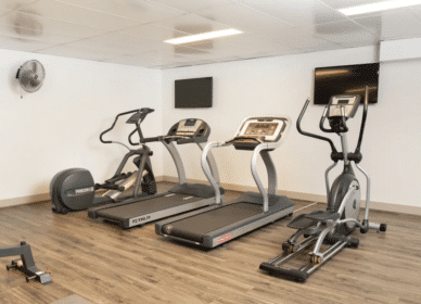 A gym room with two treadmills, an exercise bike, and an elliptical machine on a wooden floor, with two wall-mounted TVs and a fan.