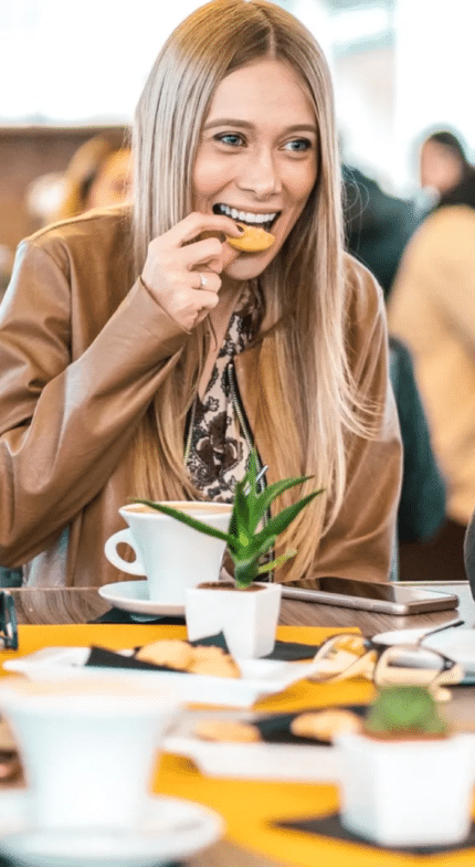 Five people sit around a table at a cafe, smiling, drinking coffee, and eating snacks. There are cups, plates, and small plants on the table.
